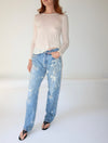 A model wearing a cream-colored long-sleeve knit top and light blue acid-wash jeans with white paint splatter details, paired with black sandals, against a plain white studio background.