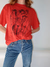 A person wearing a bright red graphic t-shirt with a black line art print of classical figures, paired with blue jeans, shown from the neck down against a plain white background.