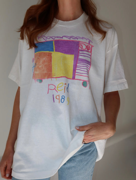 A person wearing an oversized white graphic T-shirt with a colorful abstract bus design and the text 'Reiñ 1983' on the front, paired with light blue jeans against a plain light gray wall.