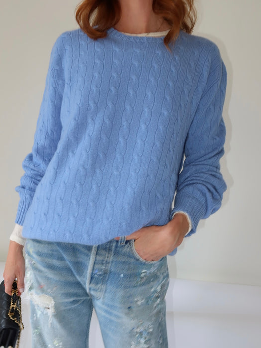 A person wearing a light blue cable-knit crewneck sweater and light-wash distressed jeans, standing against a plain light background.