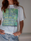 A person wearing a white graphic T-shirt featuring a Claude Monet pond scene print with a bridge and the text 'CLAUDE MONET' on the front, paired with light blue jeans against a plain light background.