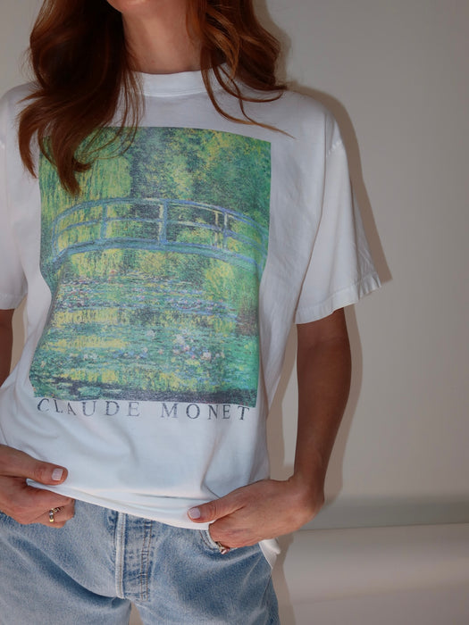 A person wearing a white graphic T-shirt featuring a Claude Monet pond scene print with a bridge and the text 'CLAUDE MONET' on the front, paired with light blue jeans against a plain light background.